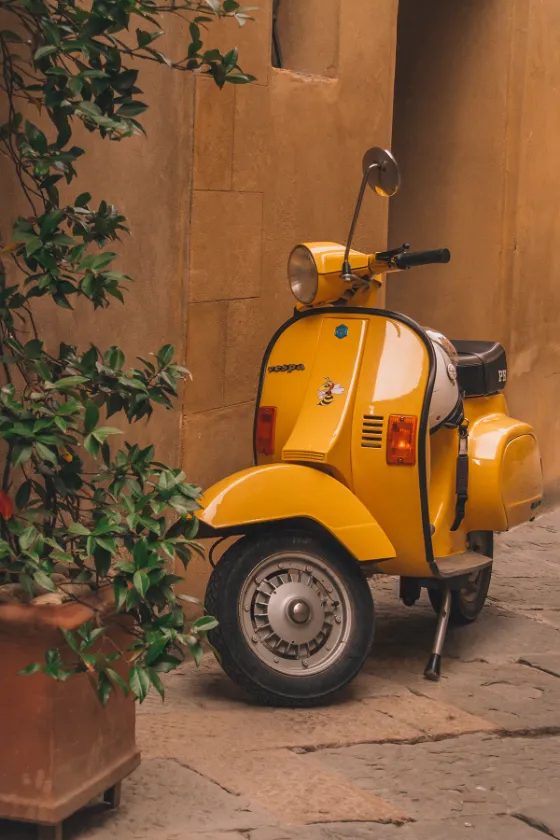 Yellow vintage scooter parked in a warm alley with ochre stucco walls, green plants, and stone pavement.