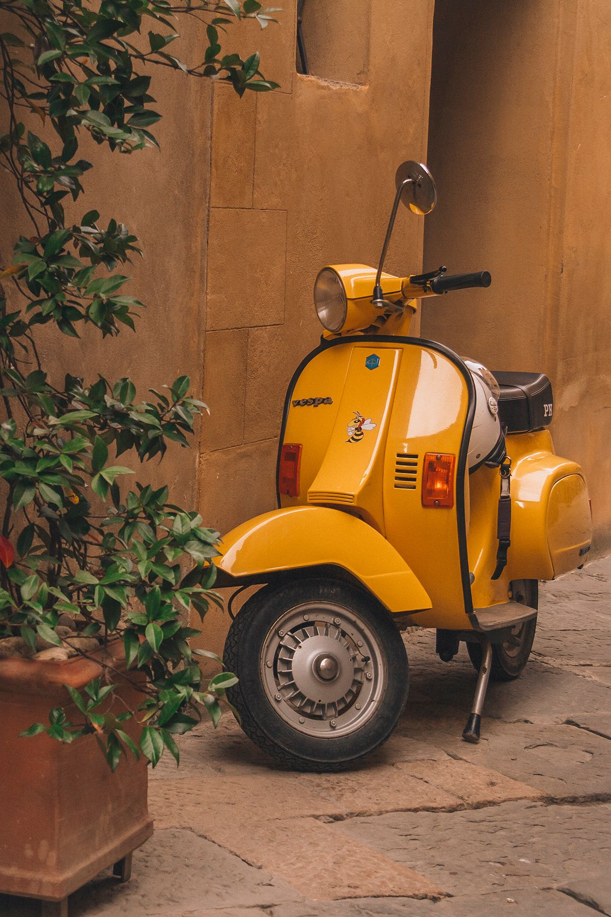 Yellow vintage scooter parked in a warm alley with ochre stucco walls, green plants, and stone pavement.