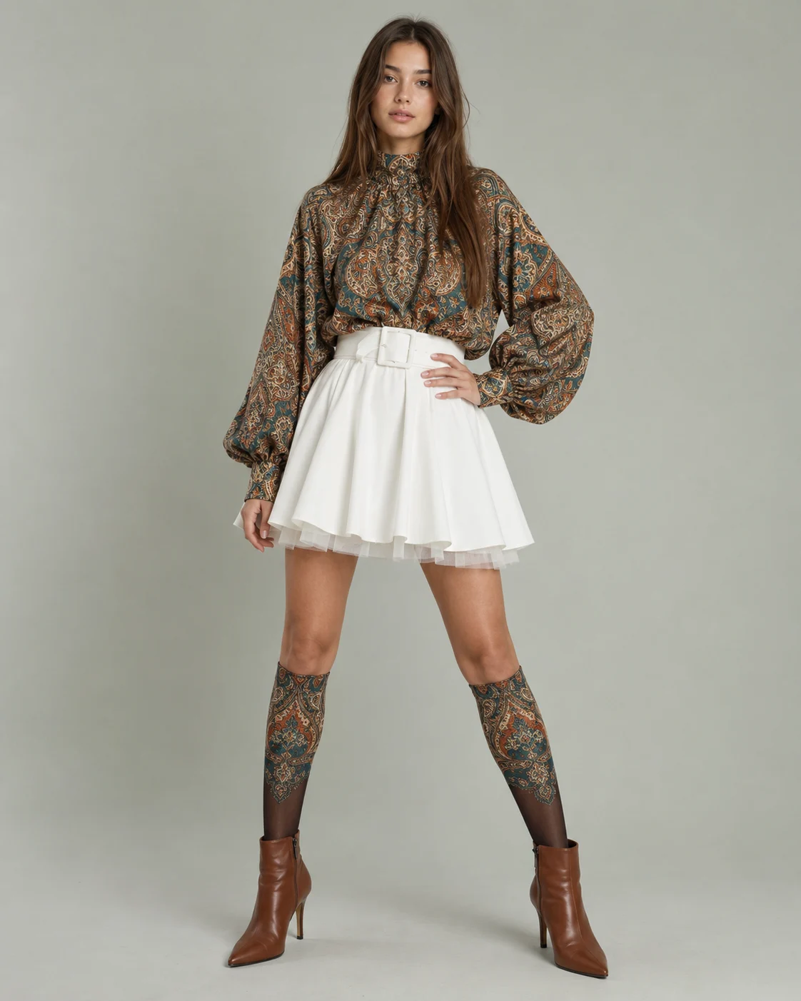Young woman in an ornate patterned high-neck blouse, white flared mini, matching patterned knee-high socks, and brown pointed ankle boots in a clean studio setting.